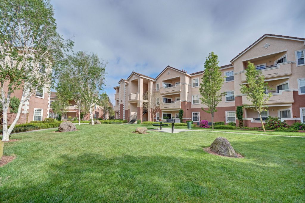 Grass courtyard between residential buildings with with lush green landscaping, picnic benches, and rock features.