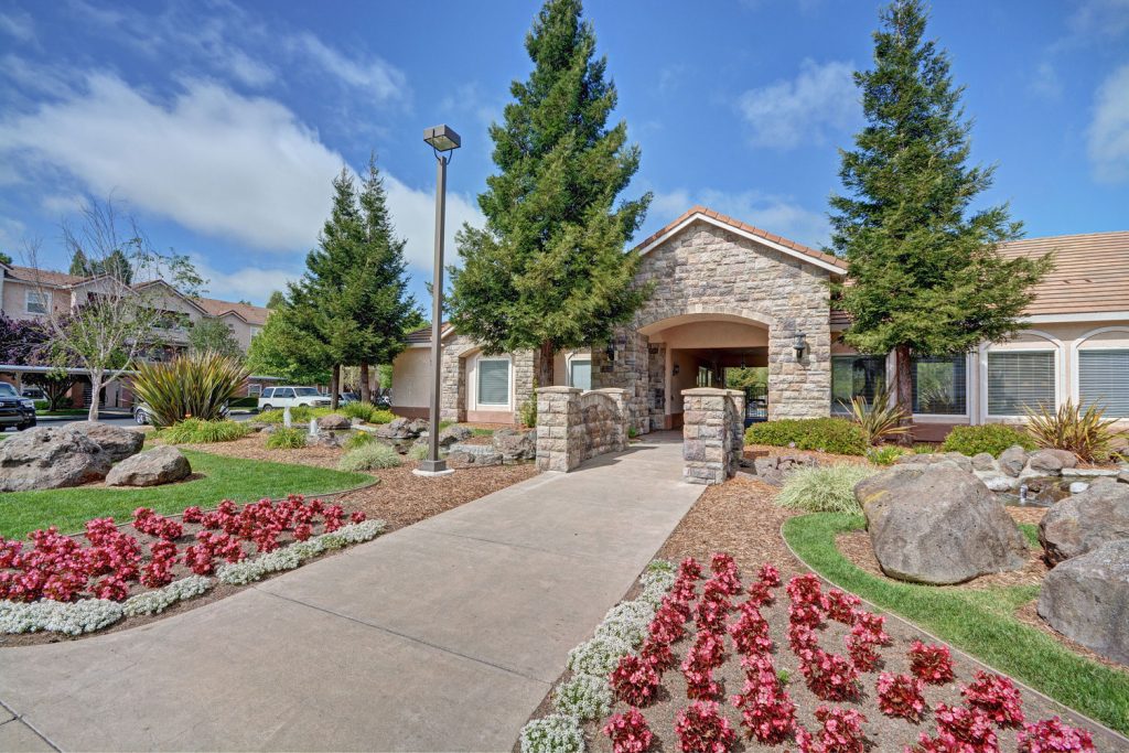 Clubhouse entrance with flowers, green landscaping, rock and water feature, and a stone bridge over water.