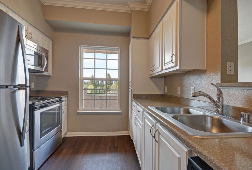 Parallel, two-wall kitchen with window, wood flooring, stainless steel appliances, two-basin sink, white cabinets, and dark countertops.