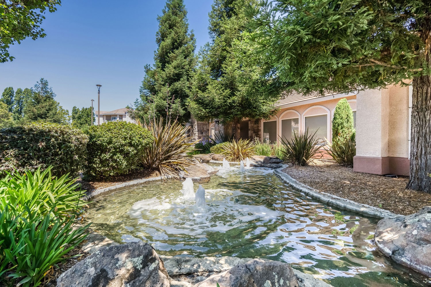 Outdoor community water feature with fountains and lush green landscaping and rocks.