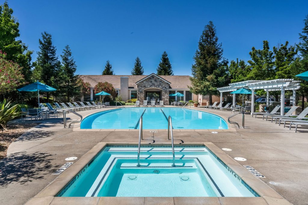 Swimming pool and spa courtyard behind the resident clubhouse with chaise lounge chairs, dining tables with umbrella coverings, and pergola shade structures.