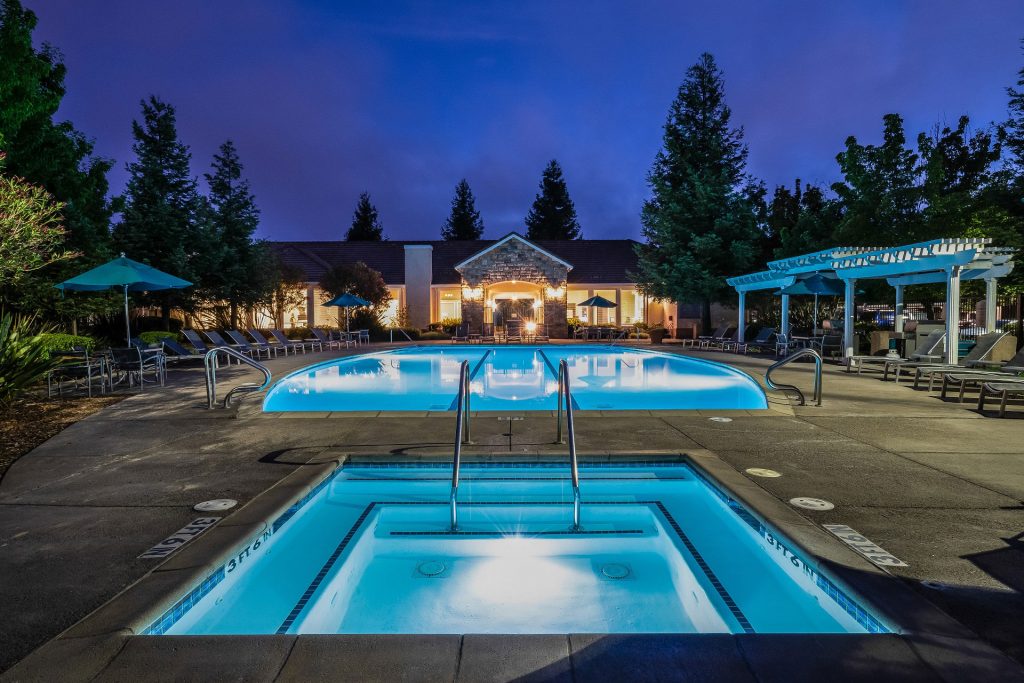 Swimming pool and spa courtyard at night with glowing lights on the resident clubhouse, chaise lounge chairs, dining tables with umbrella coverings, and pergola.