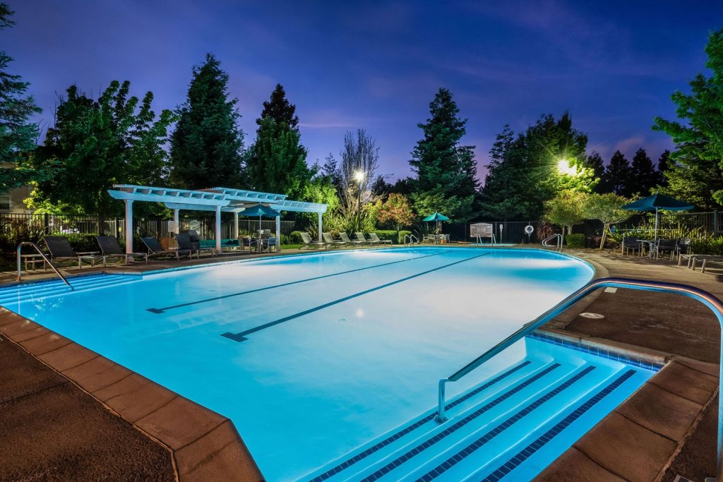 Swimming pool at night with lights, chaise lounge chairs, dining tables with umbrellas, and pergola