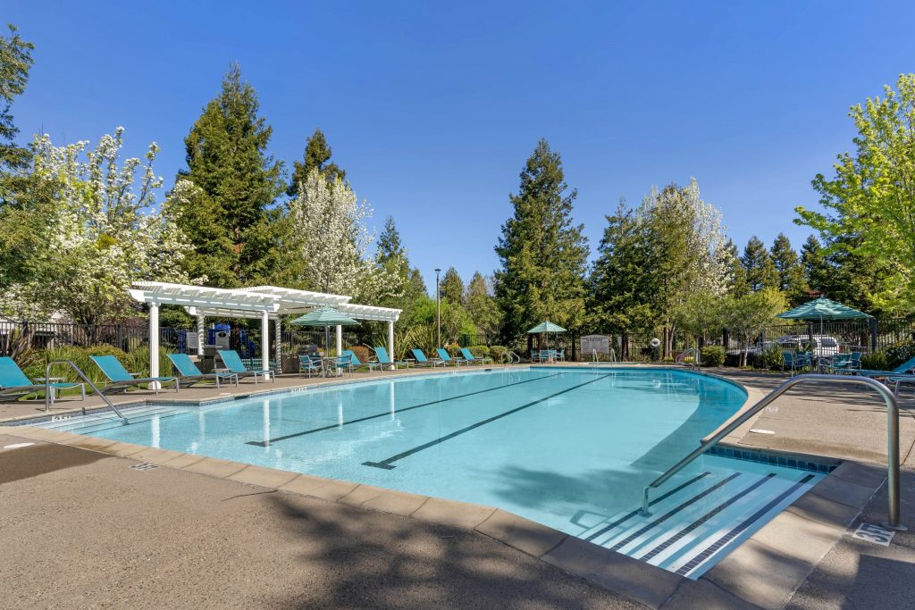 Swimming pool and spa courtyard behind the resident clubhouse with chaise lounge chairs, dining tables with umbrella coverings, and pergola shade structures.