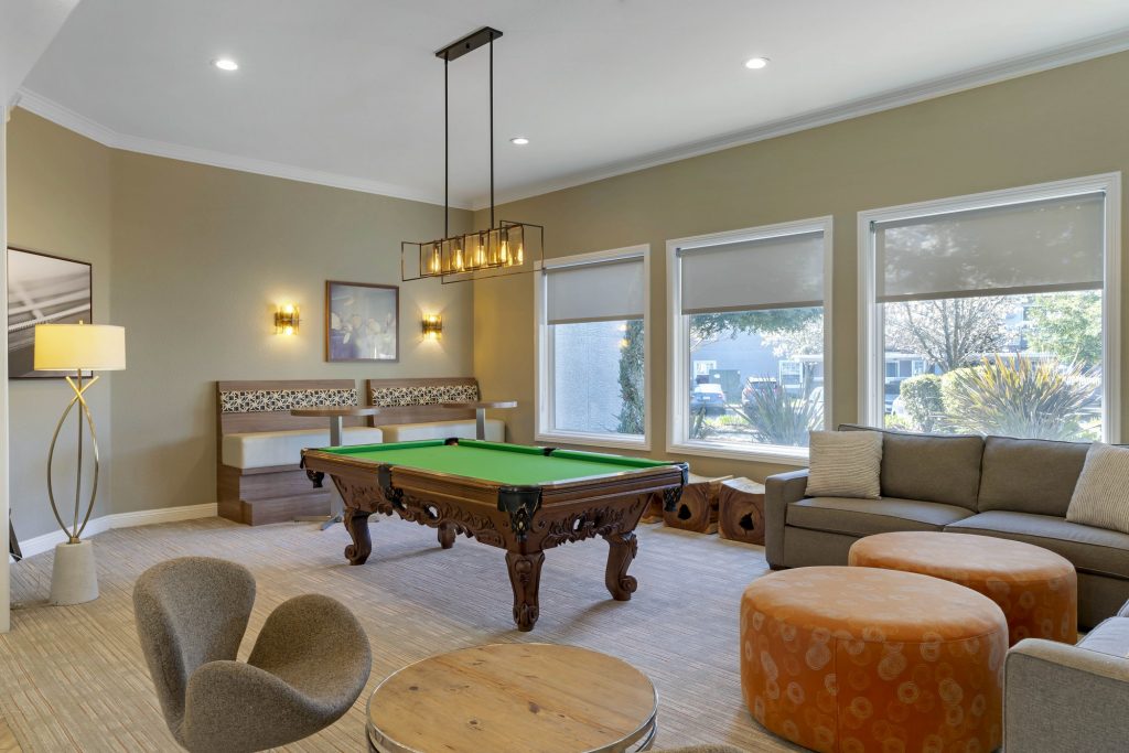 Lounge and billiards room with comfortable couches, chairs, ottomans, pool table, wall mounted TV above a fireplace, and hanging pendant lights.