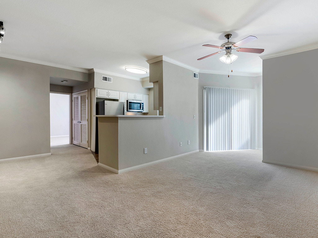 Living room area with carpet flooring, white walls, and ceiling fans, with kitchen and stainless steel appliances